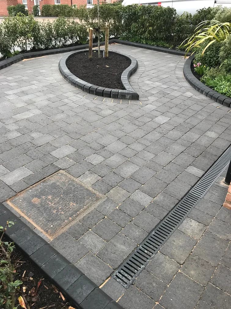Block paved driveway with curved raised borders, dark edging stones, drainage channel and planted tree.