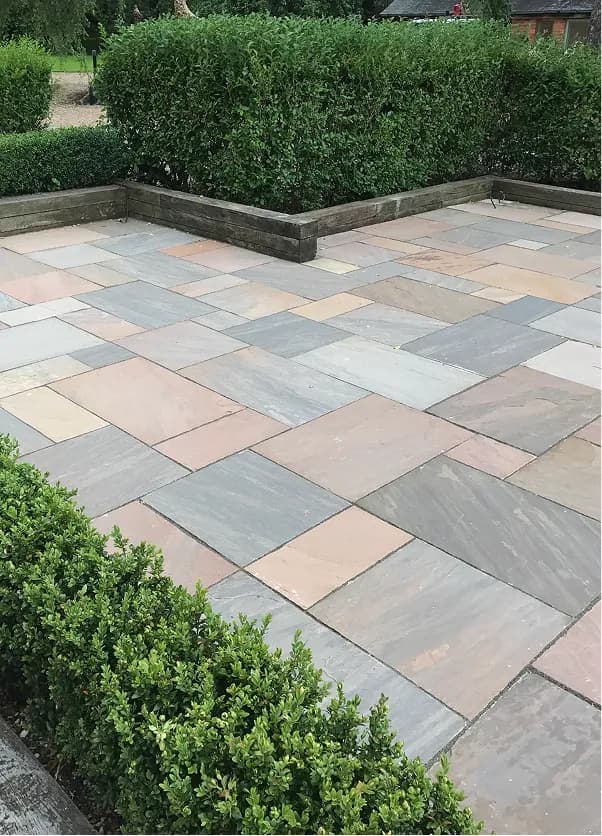 Mixed-size natural stone patio with grey and buff paving slabs, raised timber edging and garden hedging.