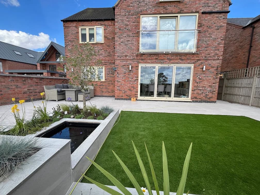 Modern back garden with artificial lawn, raised pond, porcelain patio and contemporary planting beds.